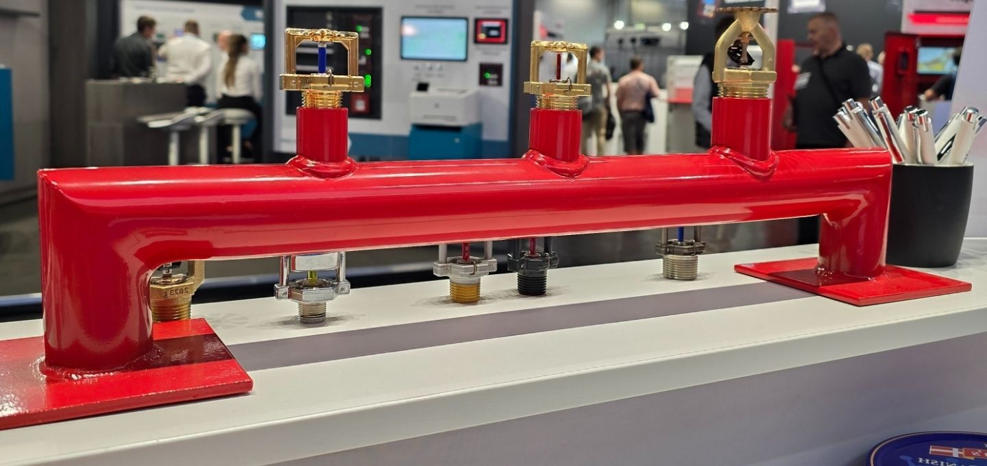Rotes Ausstellungsstück mit verschiedenen Sprinklerköpfen auf einer Brandschutzmesse, umgeben von Messeständen und Besuchern.