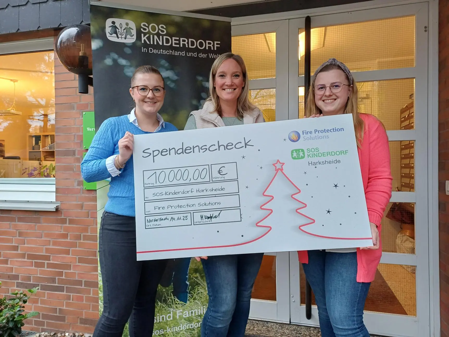 Spendenscheck über 10.000 Euro für SOS-Kinderdorf Harksheide, überreicht von Mitarbeiterinnen der Fire Protection Solutions vor dem Eingang des Kinderdorfs