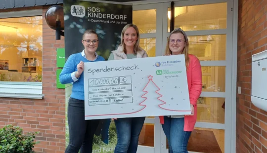 Spendenscheck über 10.000 Euro für SOS-Kinderdorf Harksheide, überreicht von Mitarbeiterinnen der Fire Protection Solutions vor dem Eingang des Kinderdorfs