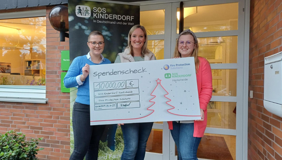Spendenscheck über 10.000 Euro für SOS-Kinderdorf Harksheide, überreicht von Mitarbeiterinnen der Fire Protection Solutions vor dem Eingang des Kinderdorfs