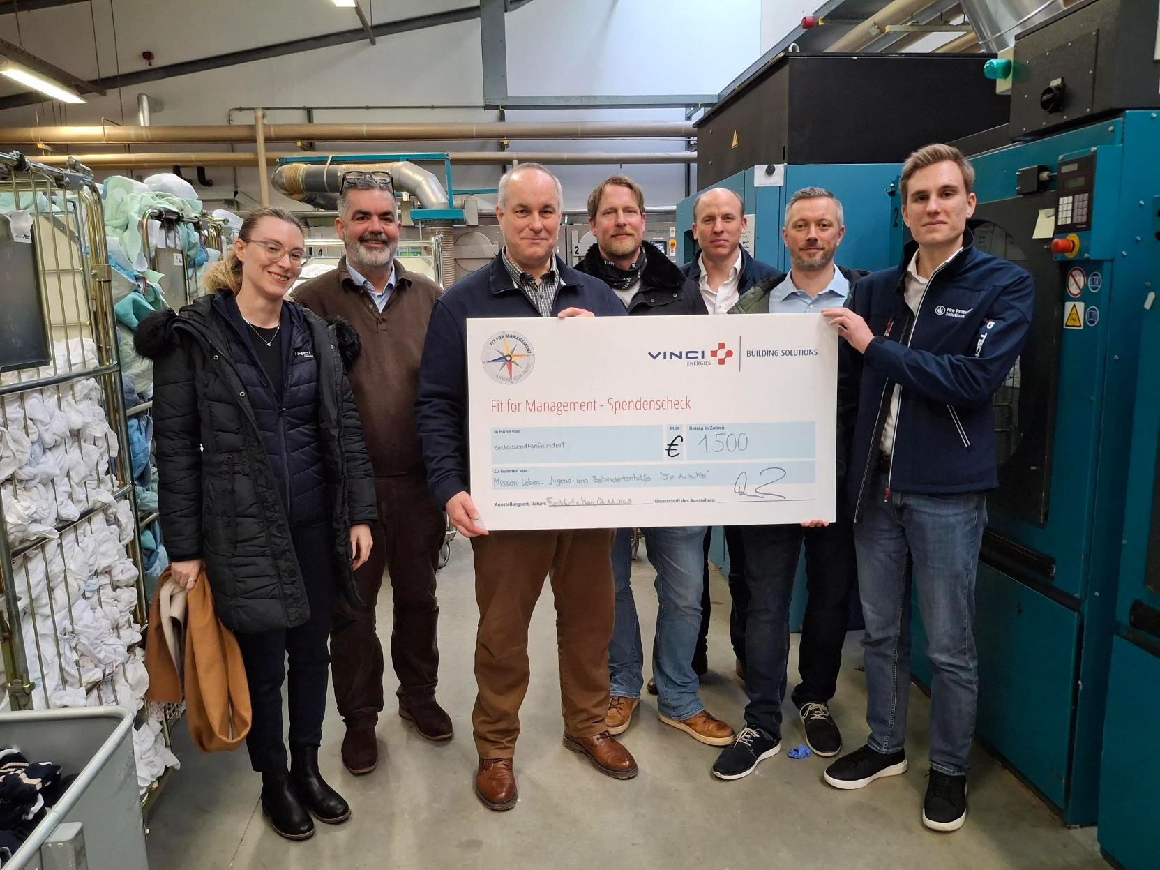 Spendenübergabe des Fit for Management Programms in der Wäscherei der Aumühle mit einem großen Spendenscheck.