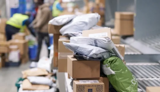 Pakete und Warensendungen auf einem Förderband im Logistikzentrum, Mitarbeitende im Hintergrund unscharf erkennbar.
